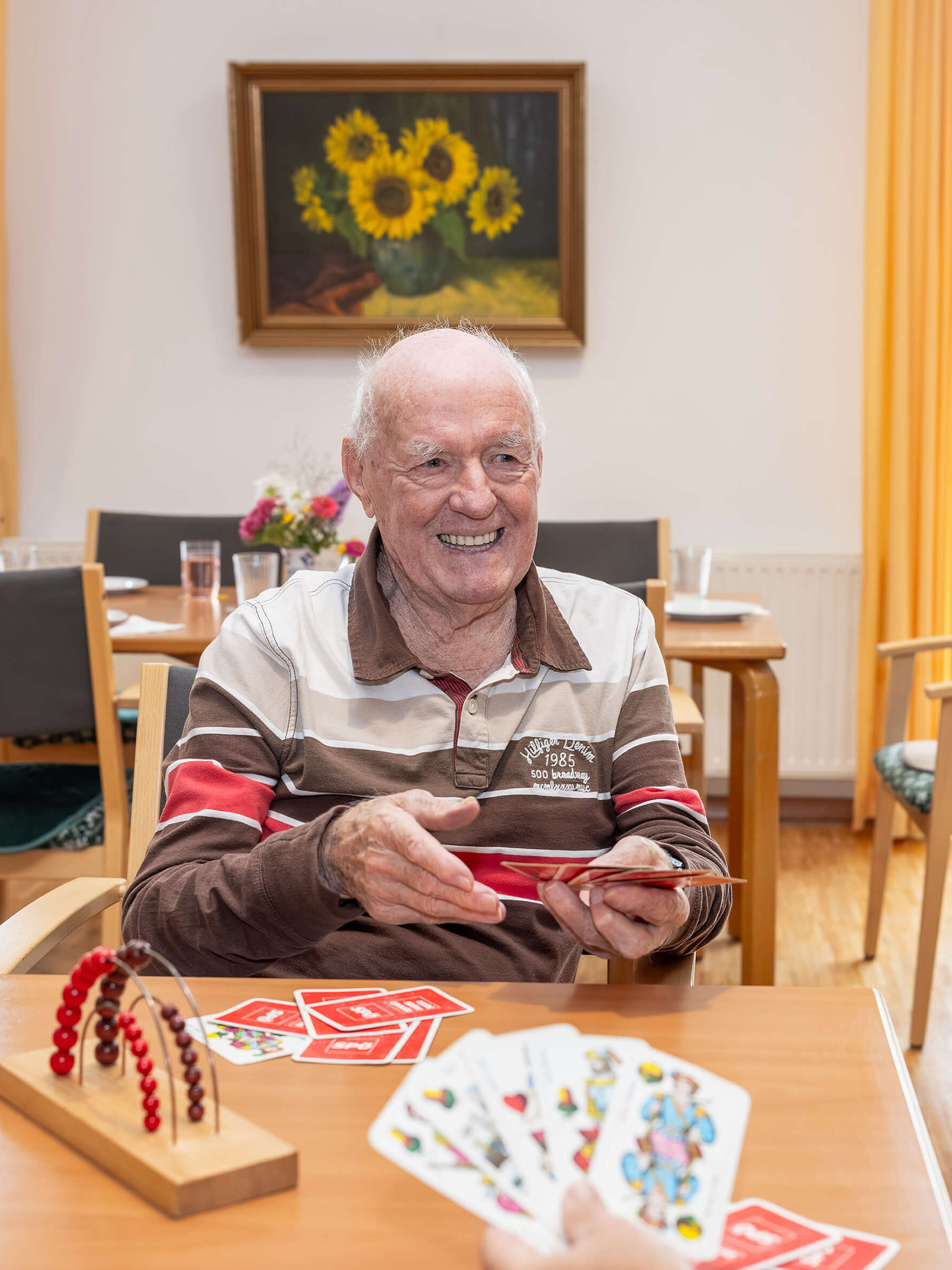 Ein Mann spielt Karten und lächelt eine Frau an, die ihm gegenübersitzt. Auf dem Tisch liegen Spielkarten.